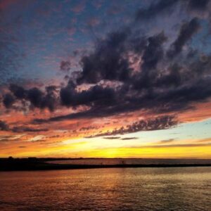 Wide-angle photo of a spectacular sunset, showcasing intense yellow, orange, and purple cloud formations above the flat horizon of the Delaware Bay.